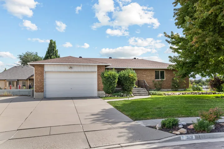 Brick exterior residence featuring an attached garage with a concrete driveway, a manicured front lawn, mature trees, and tiered concrete steps leading to the entrance