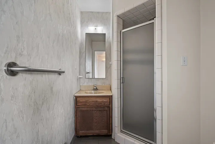Bathroom featuring a frosted glass shower enclosure with white tile surround, a wood-finish vanity with a single basin sink and light-toned countertop, and a wall-mounted mirror with overhead lighting