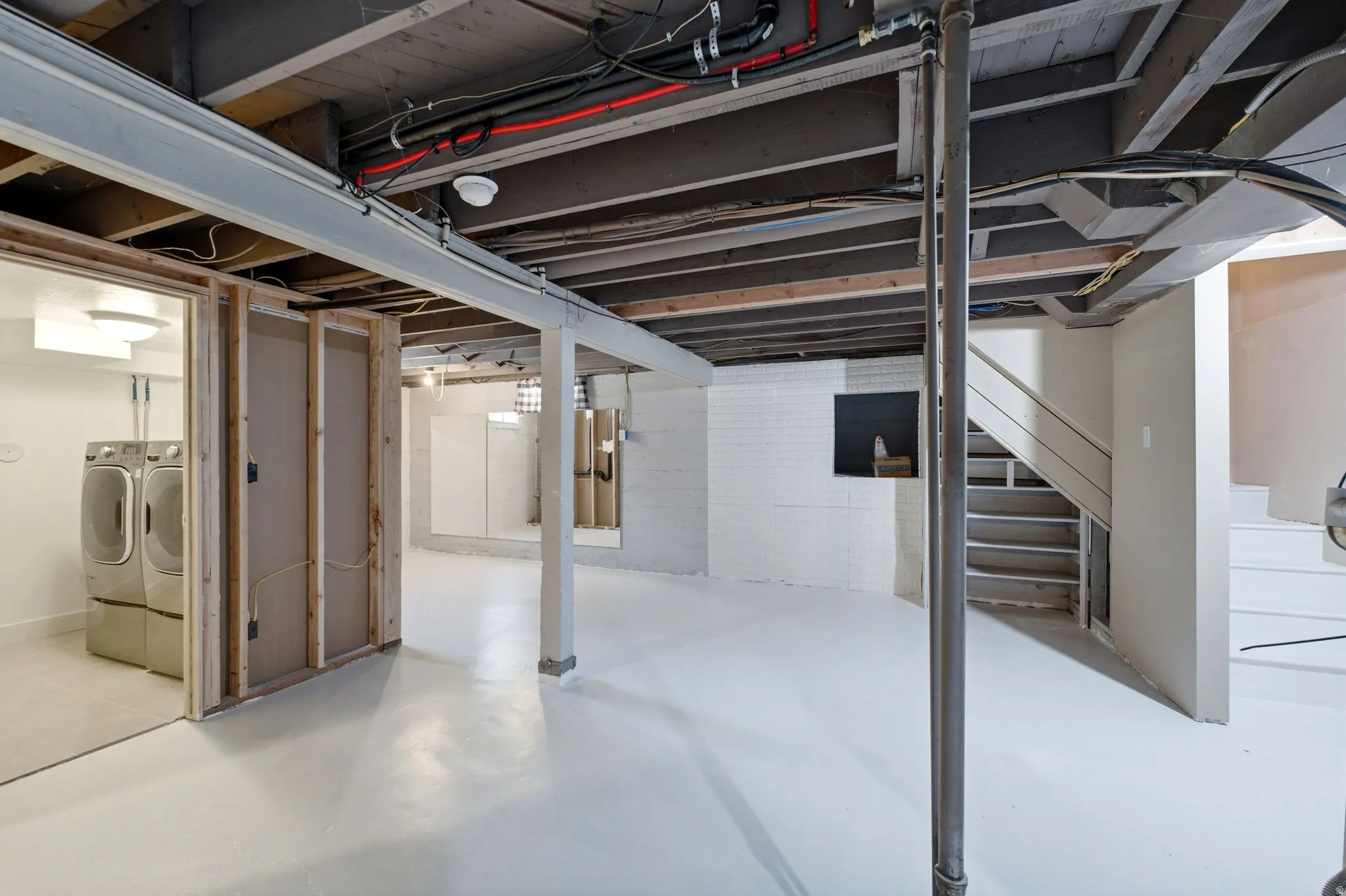 Unfinished basement space featuring exposed ceiling joists, white painted concrete flooring, and a laundry area with tile flooring