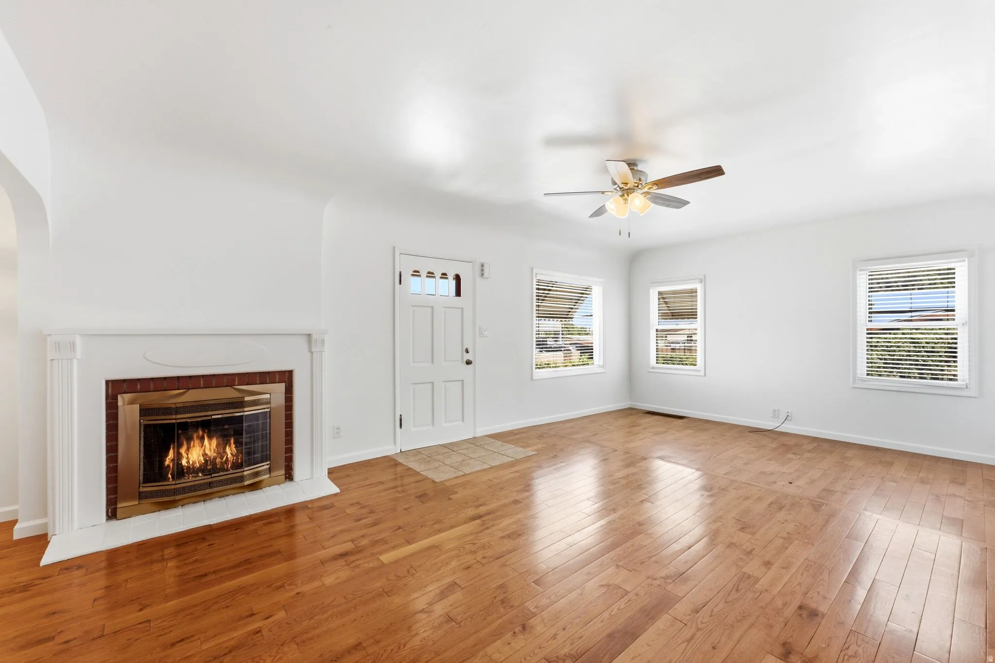 Living space featuring wood-finish flooring, a white mantel fireplace with a brass-trimmed insert, and a ceiling fan with integrated lighting