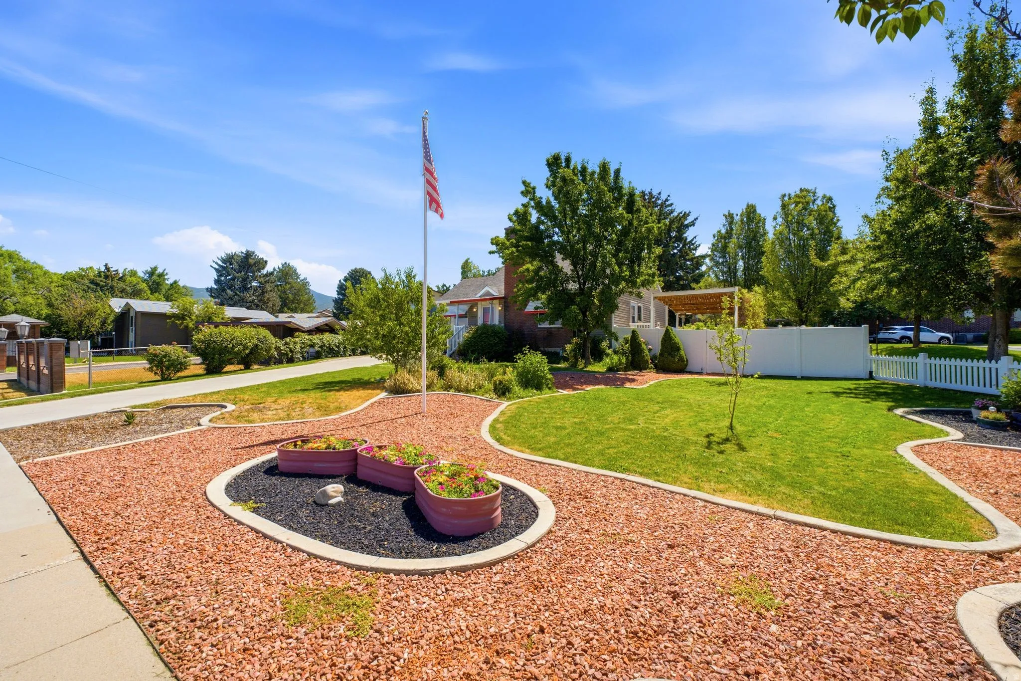 Landscaped front yard featuring a manicured lawn, decorative rock beds, a flag pole, and mature trees