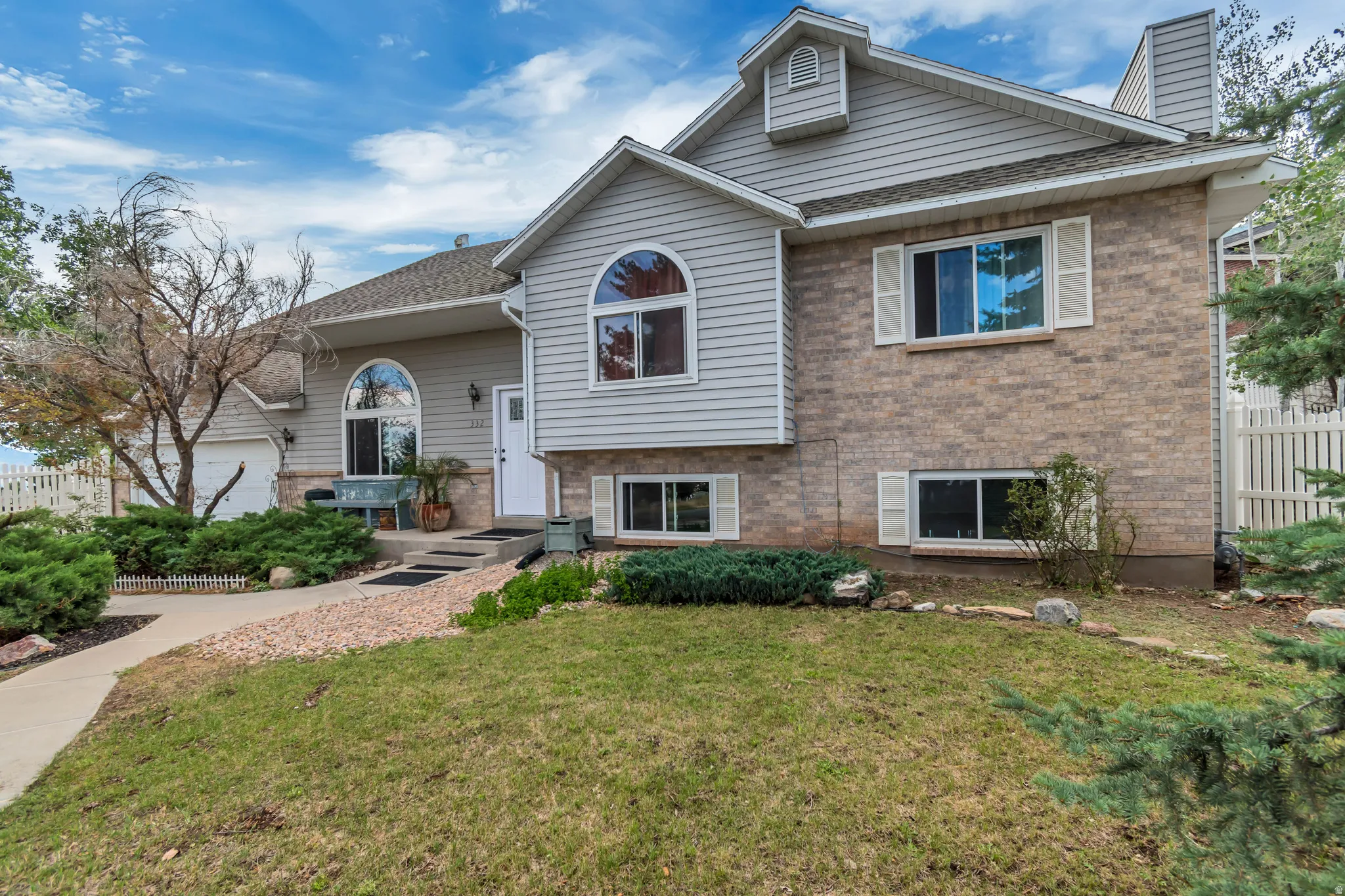 Multi-level exterior featuring brick and horizontal siding, a prominent arched window, white shutters, an attached garage, and a concrete pathway with gravel landscaping