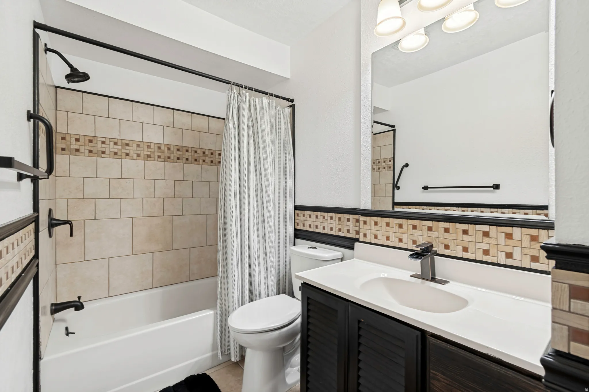 Bathroom with a tiled shower surround featuring a decorative mosaic band, a white vanity countertop with an integrated sink, a dark wood-finish vanity cabinet, a large wall-mounted mirror, and a three-light vanity fixture