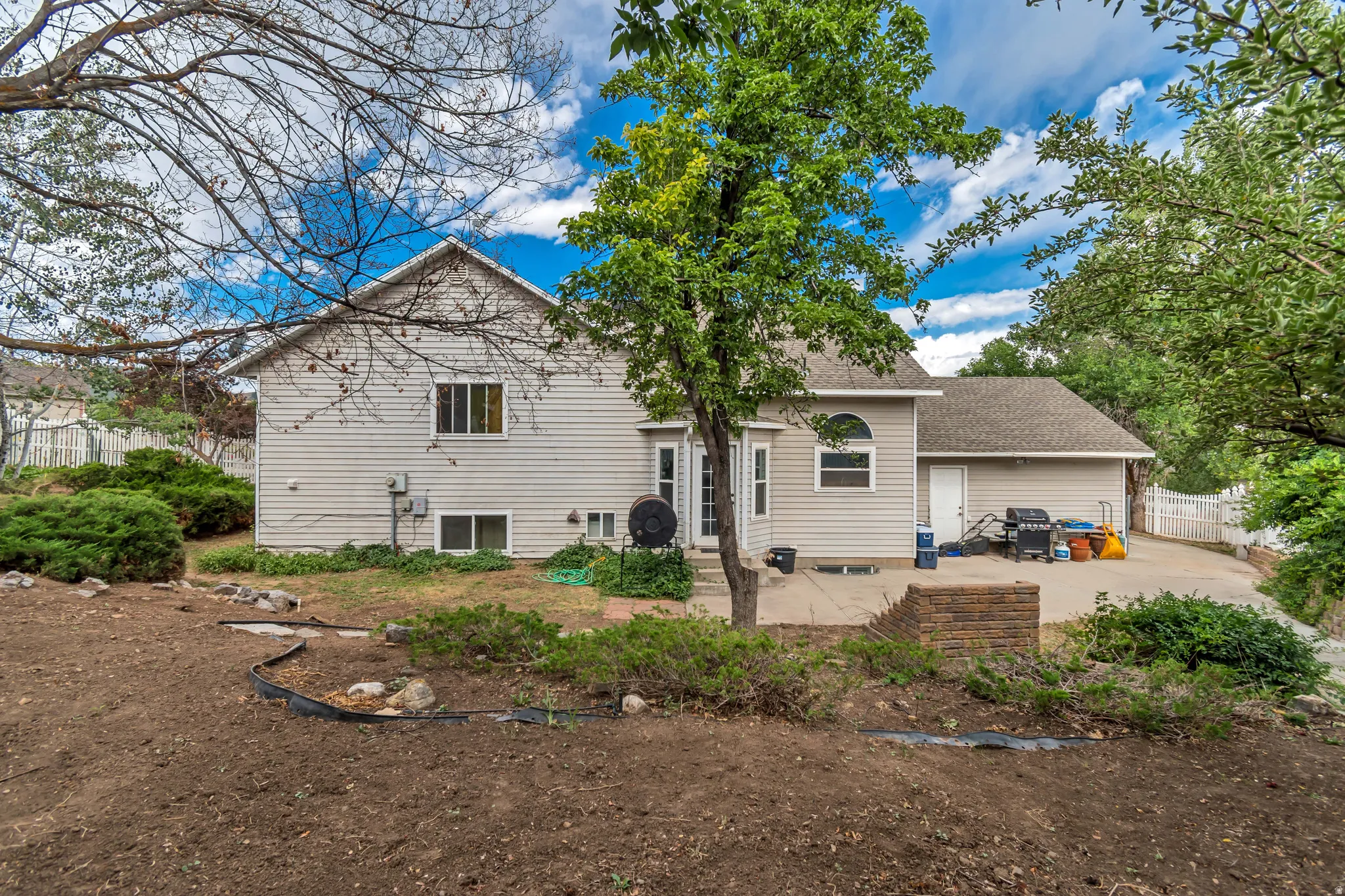 Rear exterior featuring light-colored siding, multiple windows, a stone patio, and a sloped yard with established trees and shrubs