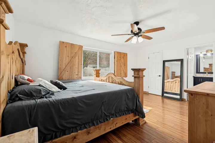 Bright bedroom featuring wood-finish flooring, a ceiling fan with integrated lighting, and natural light from a window with decorative wooden shutters