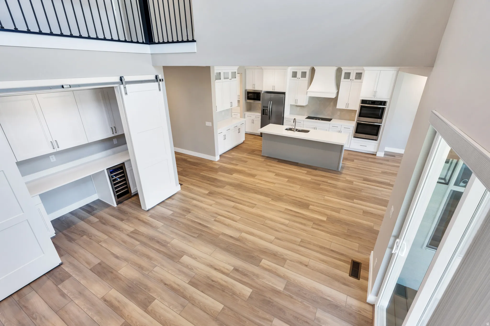 Open-concept living space featuring wood-finish flooring, a contemporary kitchen with white cabinetry and a central island, stainless steel appliances, built-in double ovens, and a dedicated workspace with a beverage refrigerator concealed by sliding barn