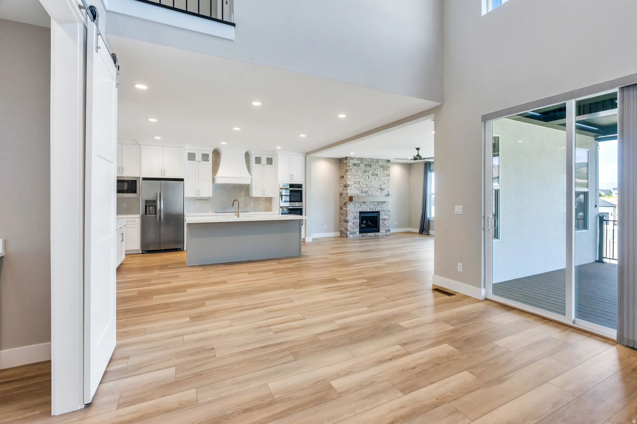 Open-concept living space featuring wood-finish flooring, a contemporary kitchen with white cabinetry and stainless steel appliances, a stone-clad fireplace, a sliding glass door leading to a covered patio, and a barn door entry