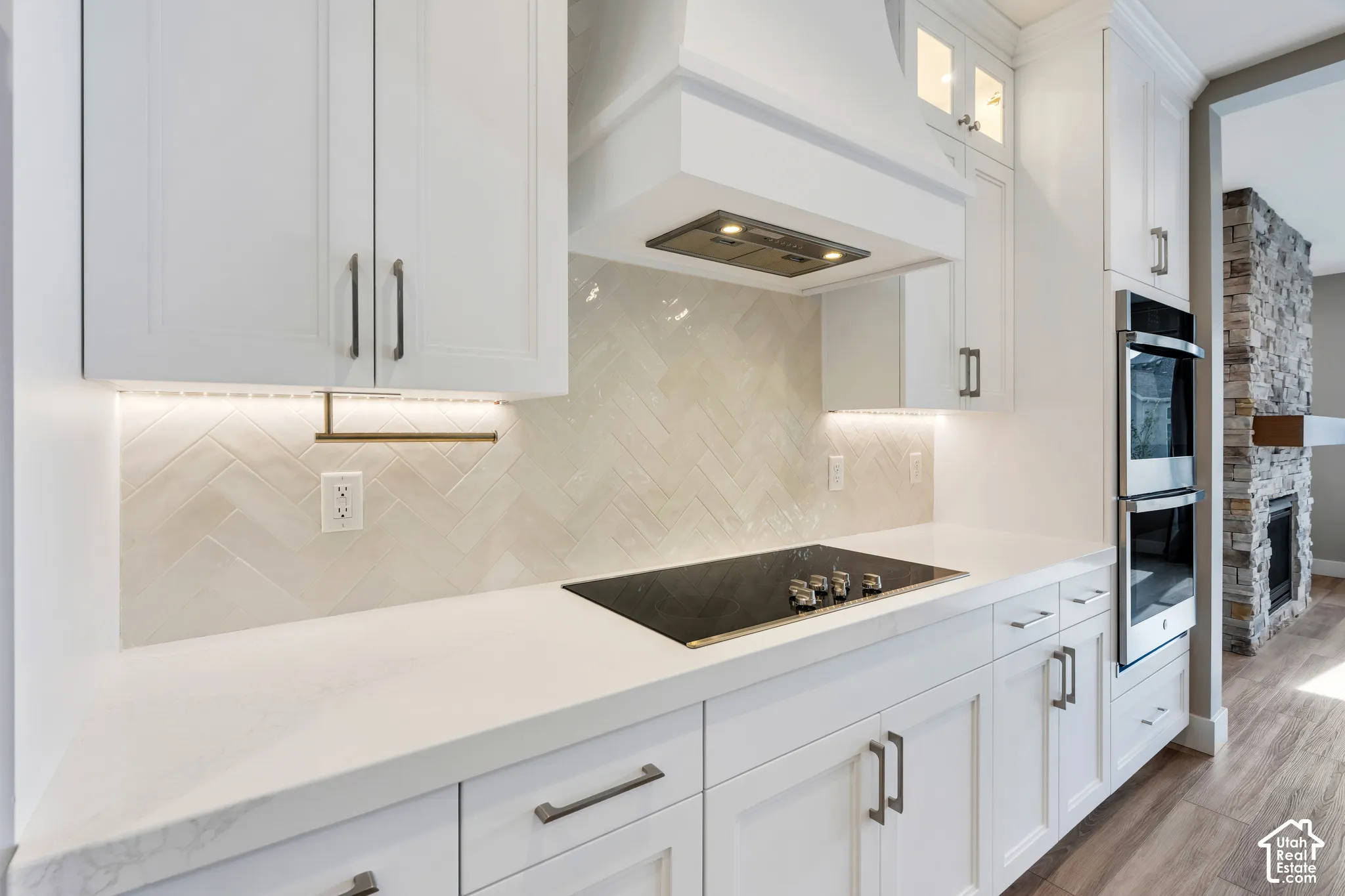 White shaker-style cabinetry with dark hardware, quartz countertops, herringbone tile backsplash with under-cabinet lighting, and a built-in double oven