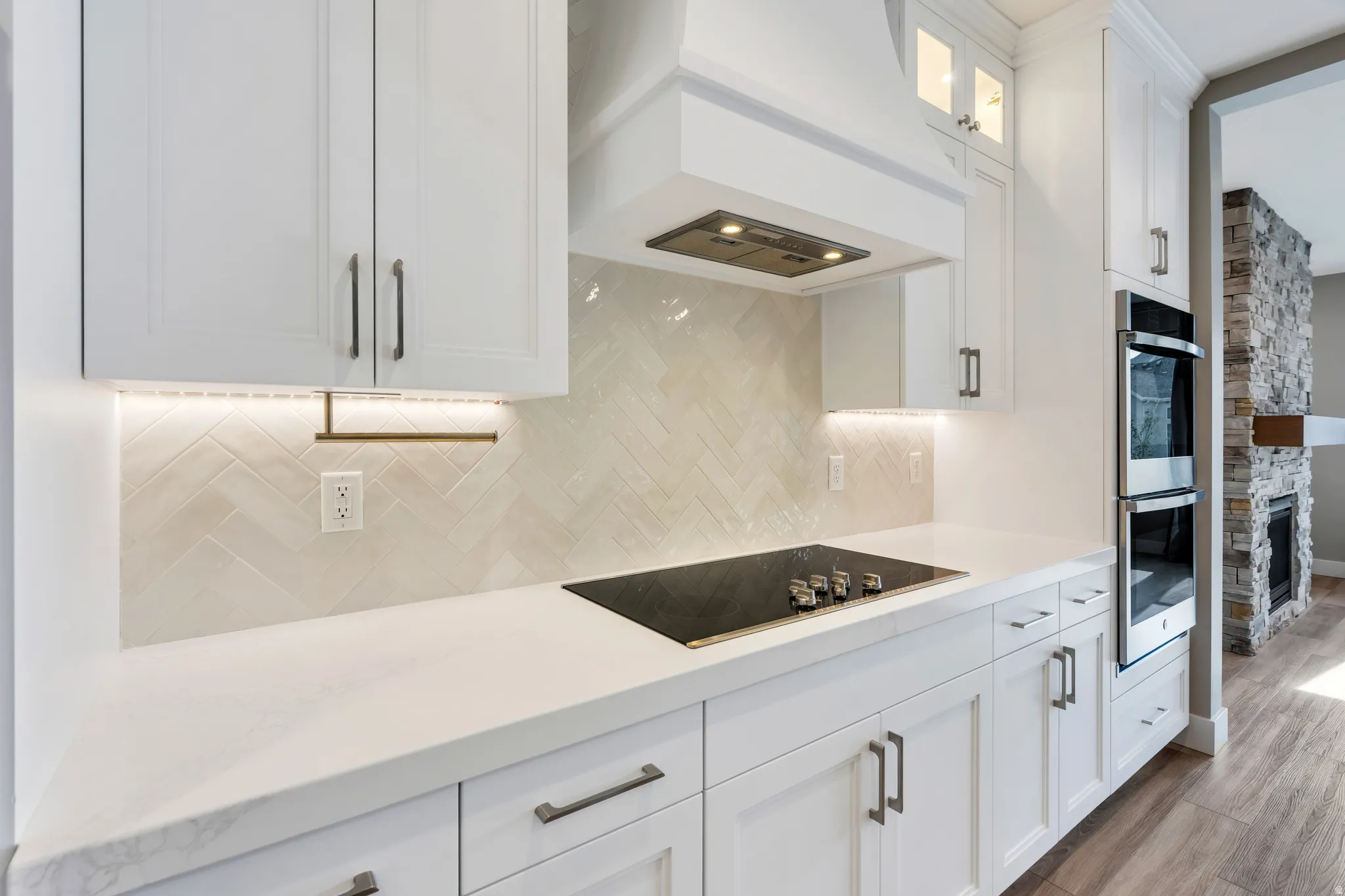 White shaker-style cabinetry with dark hardware, quartz countertops, herringbone tile backsplash with under-cabinet lighting, and a built-in double oven