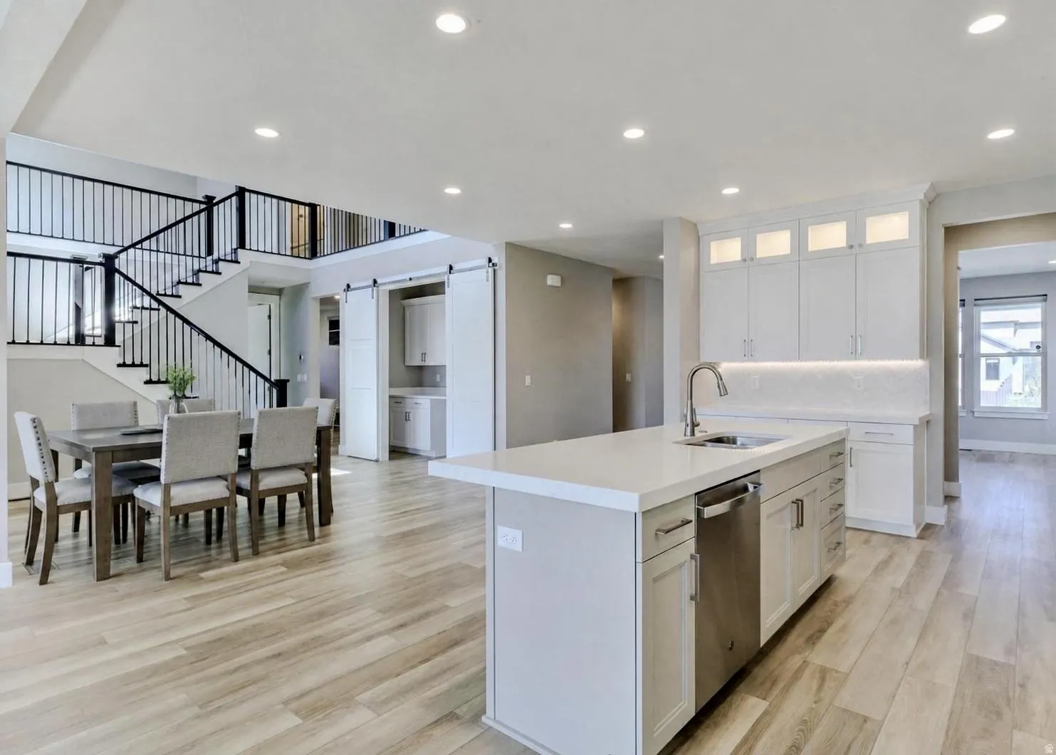 Open-concept kitchen and dining area featuring light wood-finish flooring, a kitchen island with an undermount sink and stainless steel dishwasher, white cabinetry with integrated lighting, and a contemporary staircase with black metal balusters