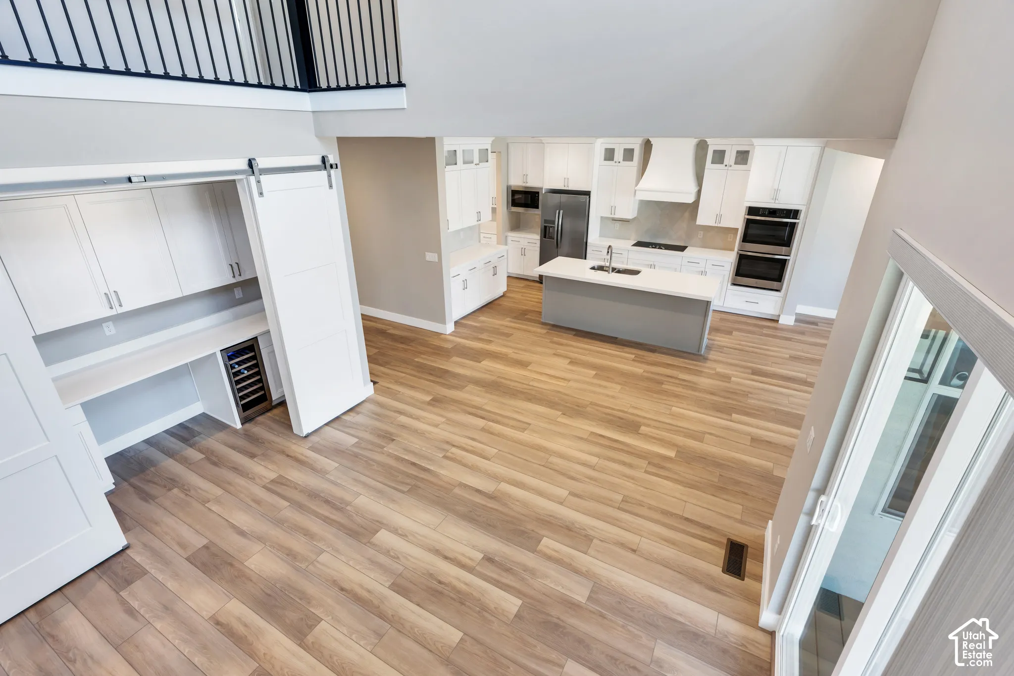 Open-concept living space featuring wood-finish flooring, a contemporary kitchen with white cabinetry and a central island, stainless steel appliances, built-in double ovens, and a dedicated workspace with a beverage refrigerator concealed by sliding barn