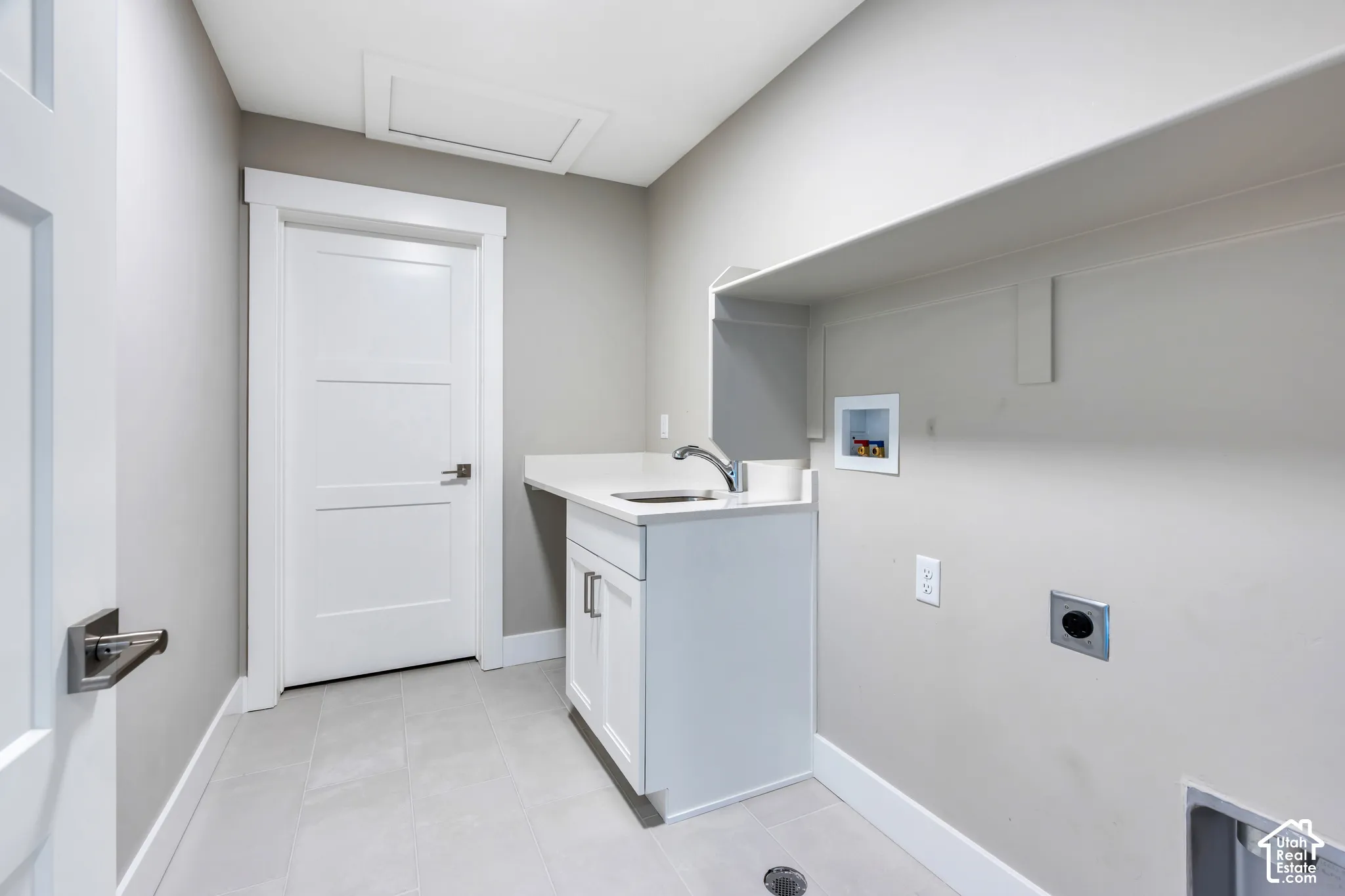 Laundry area featuring a utility sink with a chrome faucet, white cabinetry with silver hardware, an overhead storage shelf, and light-toned tile flooring