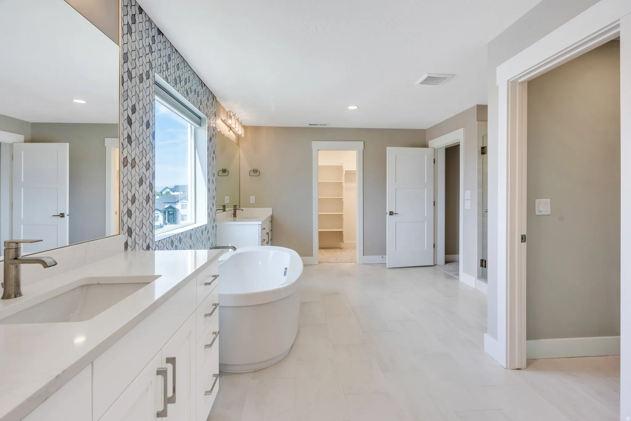 Primary bathroom with a spacious bathroom featuring a freestanding soaking tub, dual vanity with undermount sinks, patterned tile accent wall, and light-toned flooring
