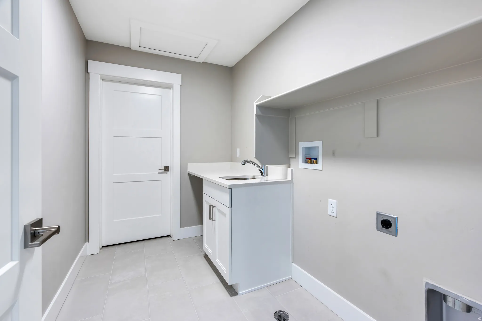 Laundry area featuring a utility sink with a chrome faucet, white cabinetry with silver hardware, an overhead storage shelf, and light-toned tile flooring