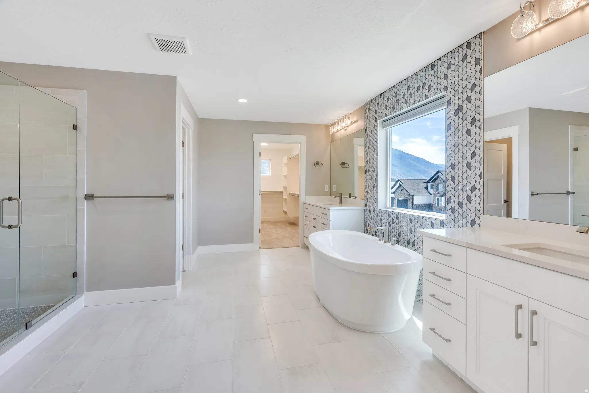 Spacious bathroom featuring a freestanding soaking tub with mosaic tile accent wall, dual vanity with white cabinetry and light countertops, a glass-enclosed shower, and large format tile flooring