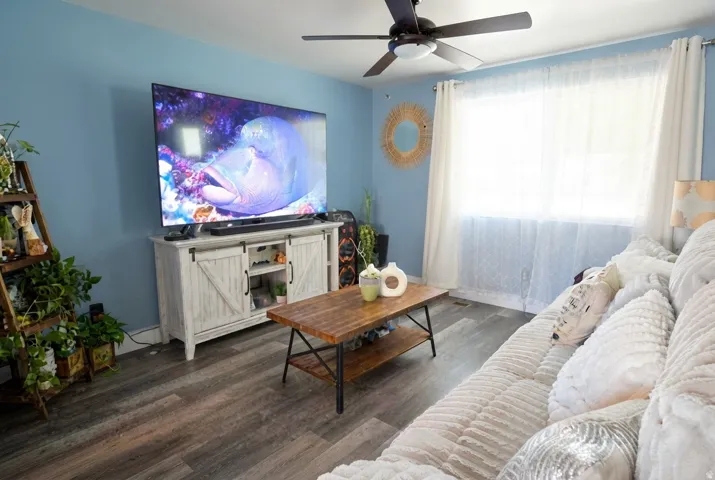 Spacious living area featuring neutral wood-finish flooring, a ceiling fan with integrated lighting, and a large window