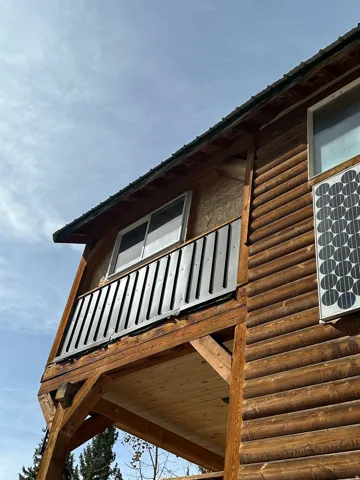 Log cabin exterior featuring a covered porch with exposed wood beams, a second-story balcony with metal railing, and a visible solar panel installation