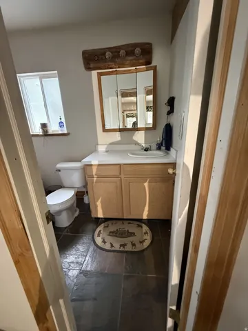 Bathroom featuring stone-finish flooring, a wood-finish vanity with a white countertop and integrated sink, a mirrored medicine cabinet, a toilet, and a window