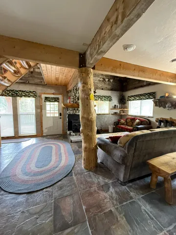 Spacious living area featuring natural stone flooring, exposed wood beams, a prominent log structural column, a stone-faced wood-burning fireplace, and multiple windows