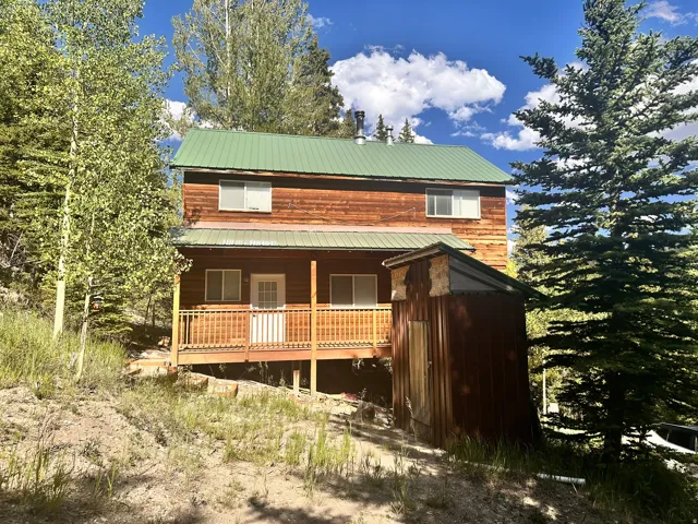 Two-story wood-sided cabin featuring a green metal roof, covered porch with wood railing, and a side outbuilding