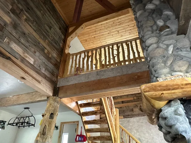 Rustic interior featuring a stone fireplace, wood-plank walls, exposed wood beams, a log staircase, and a wood branch-style railing