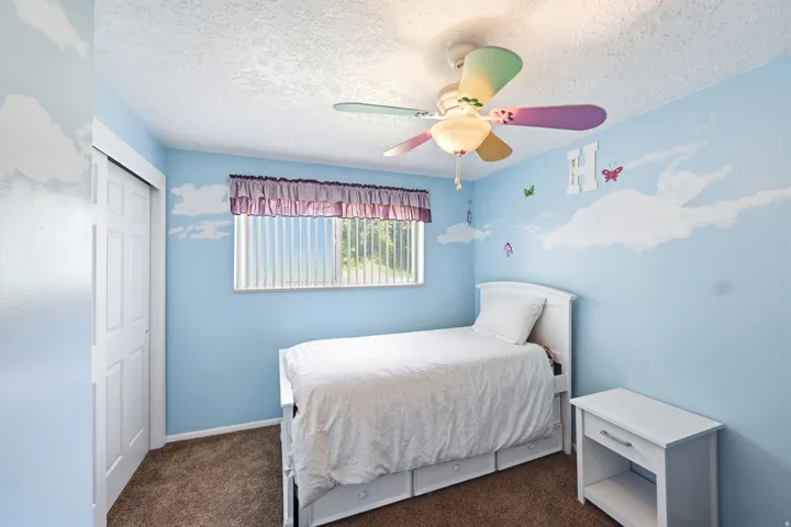 Bedroom featuring blue walls with cloud detailing, a textured ceiling, a window with vertical blinds, and a ceiling fan with multi-colored blades