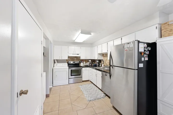 Kitchen featuring white cabinetry, tile flooring, stainless steel appliances, a tile backsplash, and an integrated laundry area with a washer and dryer