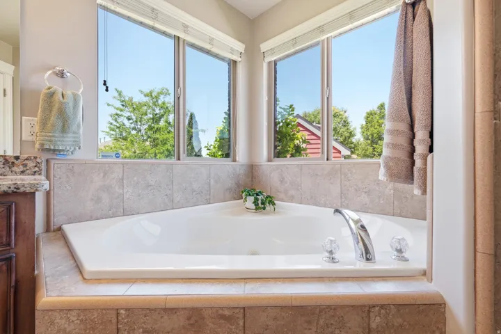 Spacious corner soaking tub featuring a chrome faucet with clear knobs, surrounded by large format tile, and framed by two corner windows with blinds