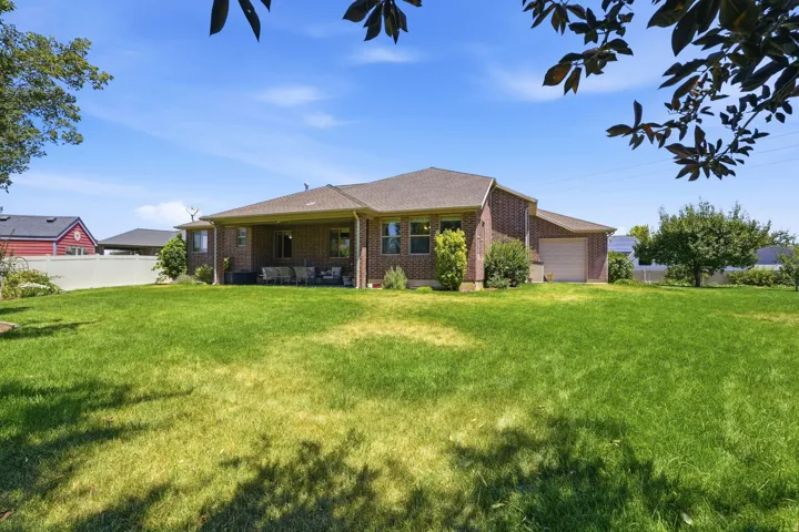 Expansive green lawn surrounds a brick residence featuring a covered patio, multiple windows with dark trim, and a detached garage with an overhead door