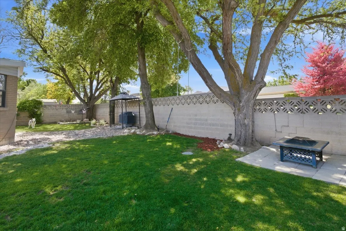Expansive backyard featuring a lush lawn, mature trees providing shade, a concrete block privacy wall with decorative elements, a paved patio area, and a dedicated fire pit space