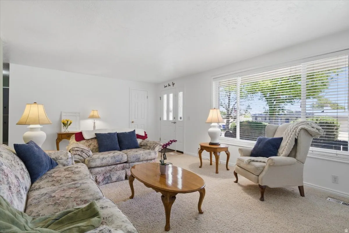 Bright living space featuring neutral carpet flooring, white walls, large window with blinds, and a white entry door with frosted glass panels
