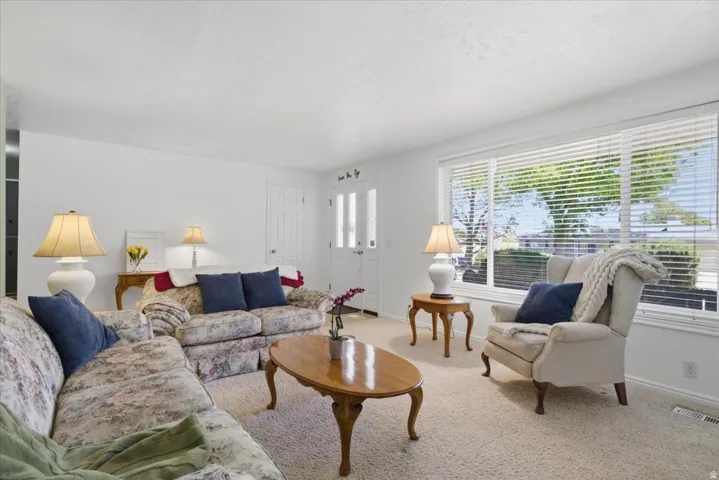 Bright living space featuring neutral carpet flooring, white walls, large window with blinds, and a white entry door with frosted glass panels