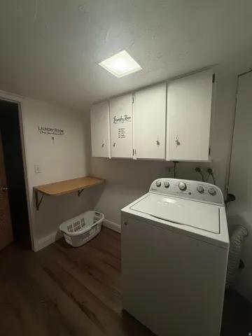 Dedicated laundry area featuring wood-finish flooring, overhead white cabinetry, a built-in folding shelf, and overhead lighting
