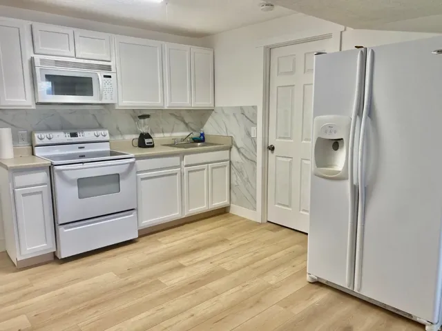 Kitchen featuring white cabinetry, light-toned countertops, a full suite of white appliances, a patterned backsplash, and wood-finish flooring