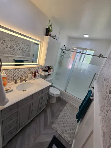 Bathroom featuring a grey vanity with a light-colored countertop, an illuminated mirror, patterned backsplash, and wood-finish flooring in a herringbone pattern
