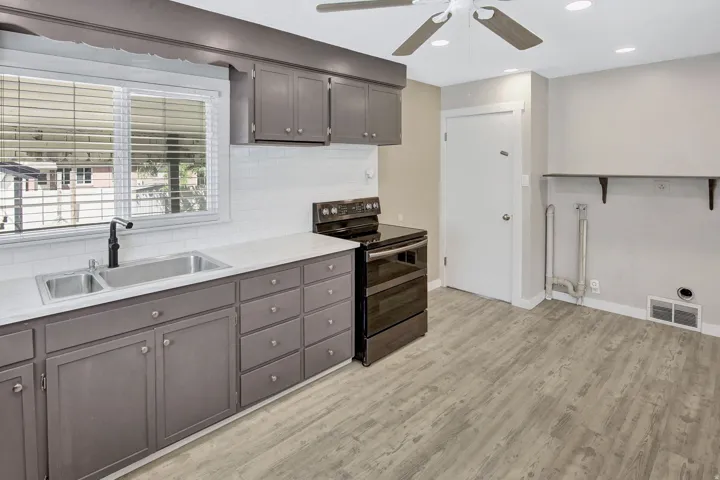 Kitchen area featuring dark gray cabinetry, a white subway tile backsplash, a stainless steel double basin sink with a matte black faucet, and light wood-finish flooring