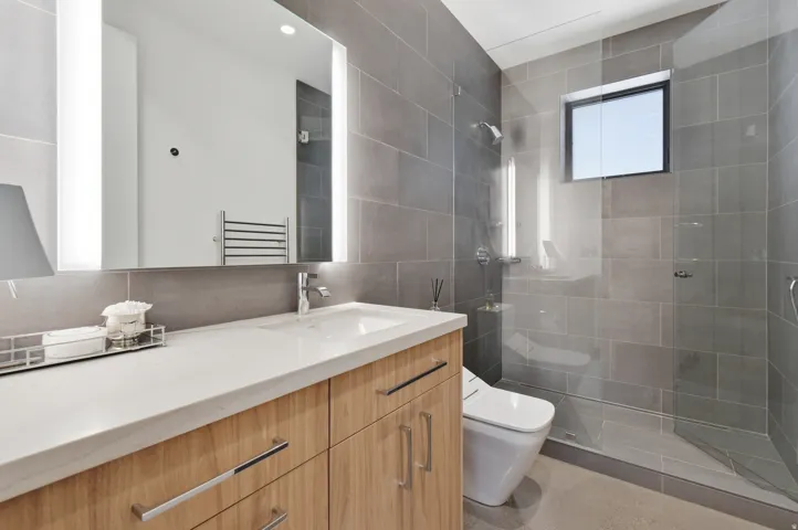 Modern bathroom featuring a wood-finish vanity with an integrated sink and light-toned countertop, a back-lit mirror, and a glass-enclosed shower with gray tiled walls