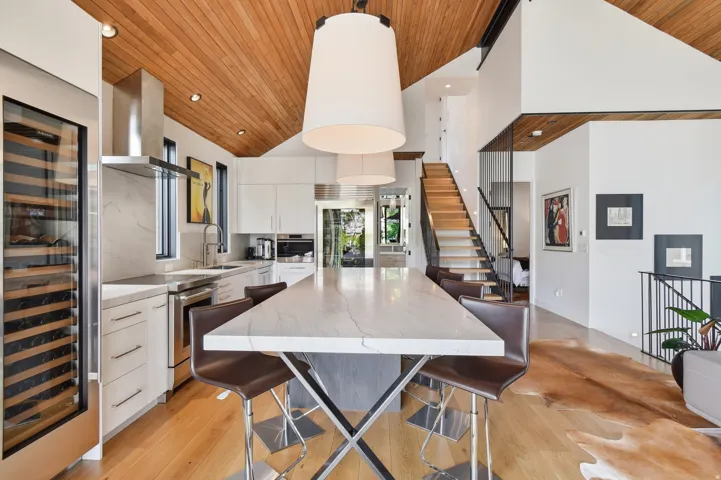 Contemporary kitchen featuring a large central island with seating, stainless steel appliances, a built-in wine cooler, wood-finish flooring, and a vaulted wood-paneled ceiling