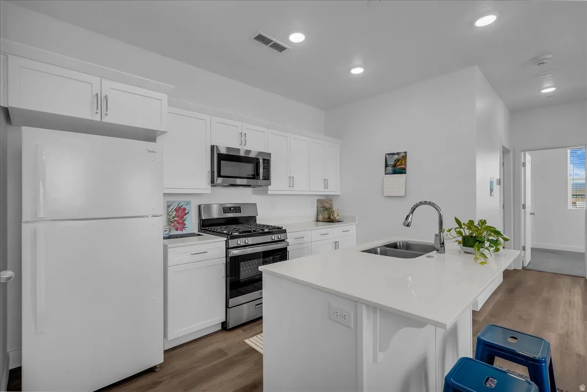 Modern kitchen featuring white cabinetry, stainless steel appliances, a center island with a double basin sink, light-toned countertops, and wood-finish flooring