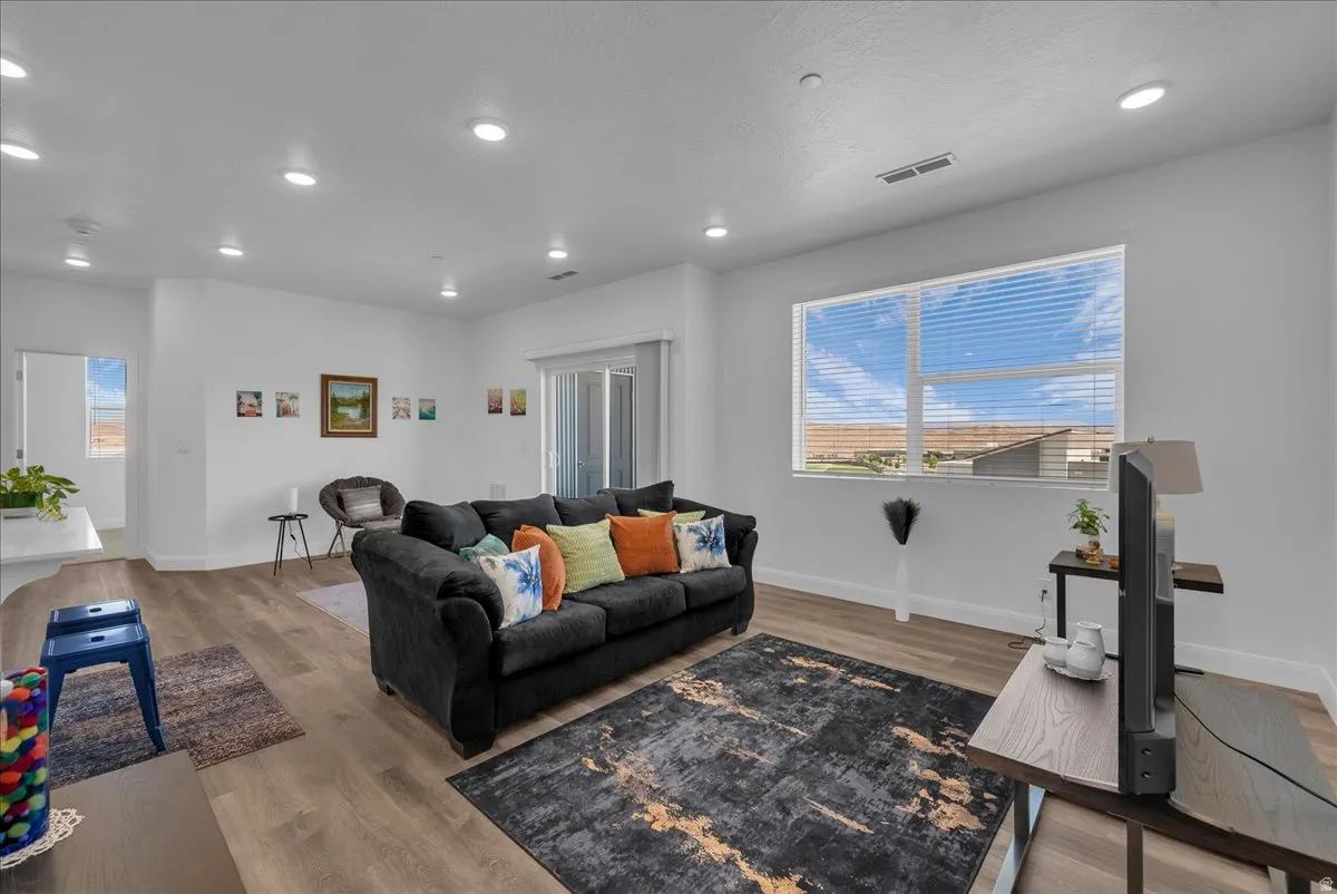 Spacious living area featuring neutral white walls, wood-finish flooring, recessed lighting, and a large window with horizontal blinds