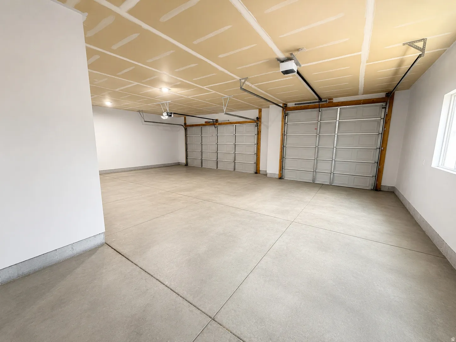 Spacious garage featuring two overhead doors with automatic openers, a concrete floor, bright overhead lighting, and a single window for natural illumination