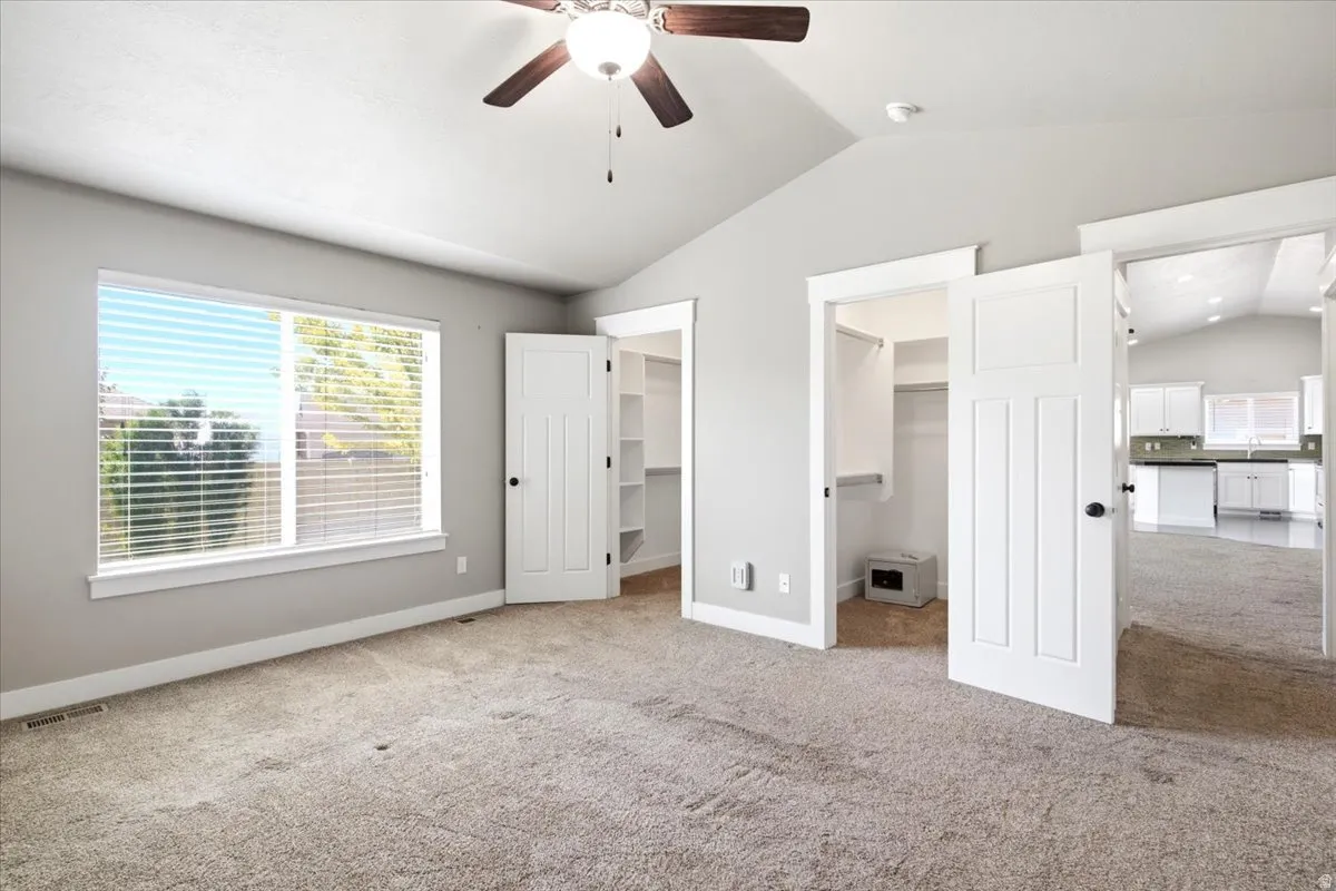 Carpeted room featuring a vaulted ceiling with a ceiling fan, a large window with blinds, and two open doorways revealing interior spaces