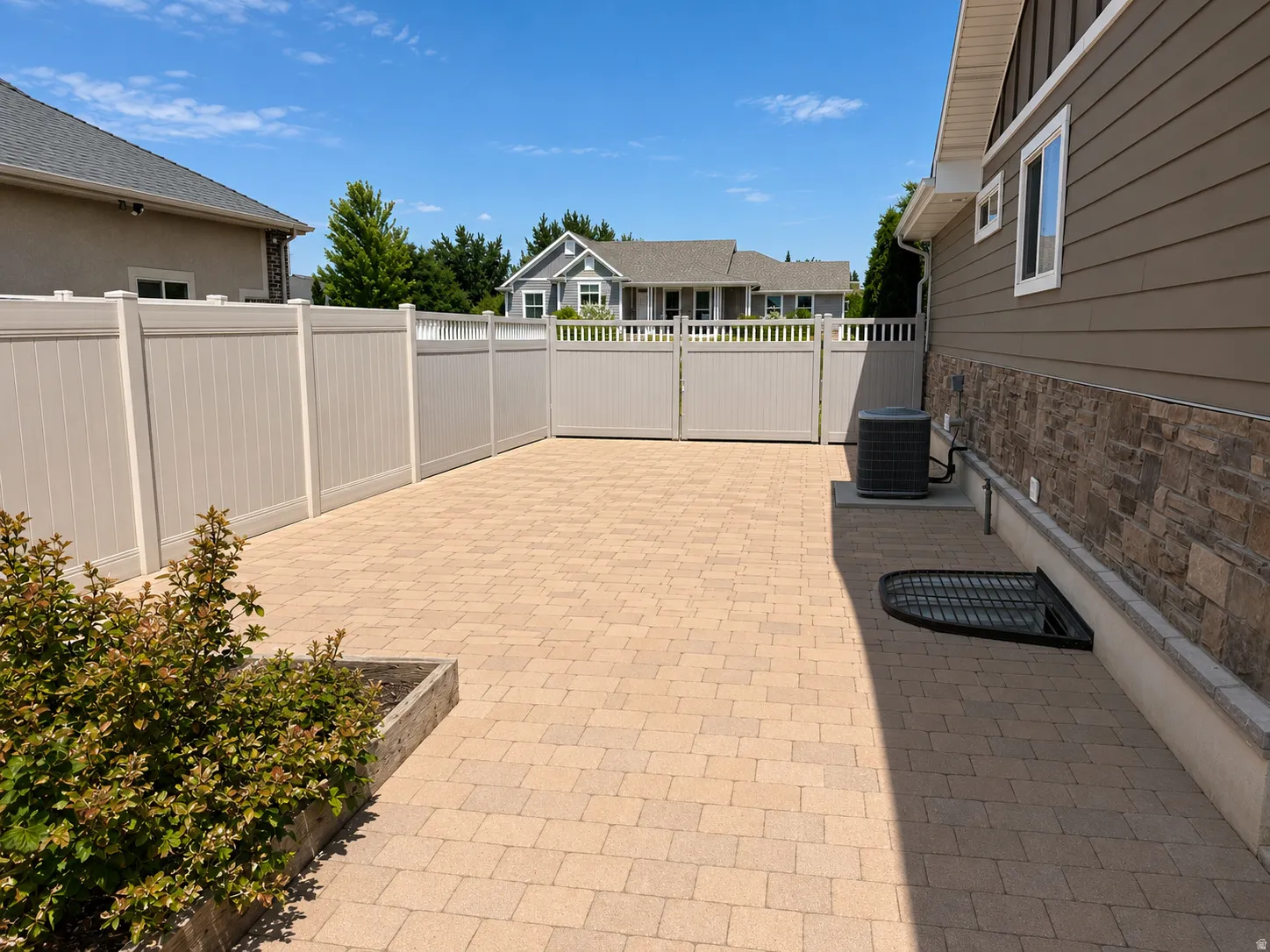 Paved outdoor area featuring light-toned paver stones, a substantial privacy fence with double gates, and a raised planter box with shrubs