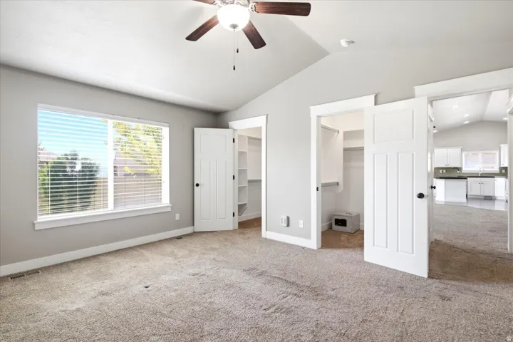 Carpeted room featuring a vaulted ceiling with a ceiling fan, a large window with blinds, and two open doorways revealing interior spaces