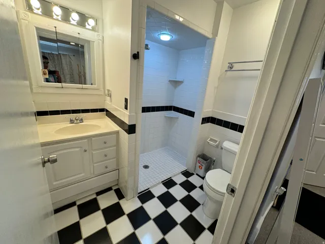Bathroom featuring a checkerboard tile floor, a white vanity with integrated sink, an illuminated medicine cabinet, a tiled walk-in shower with built-in shelving, and a toilet