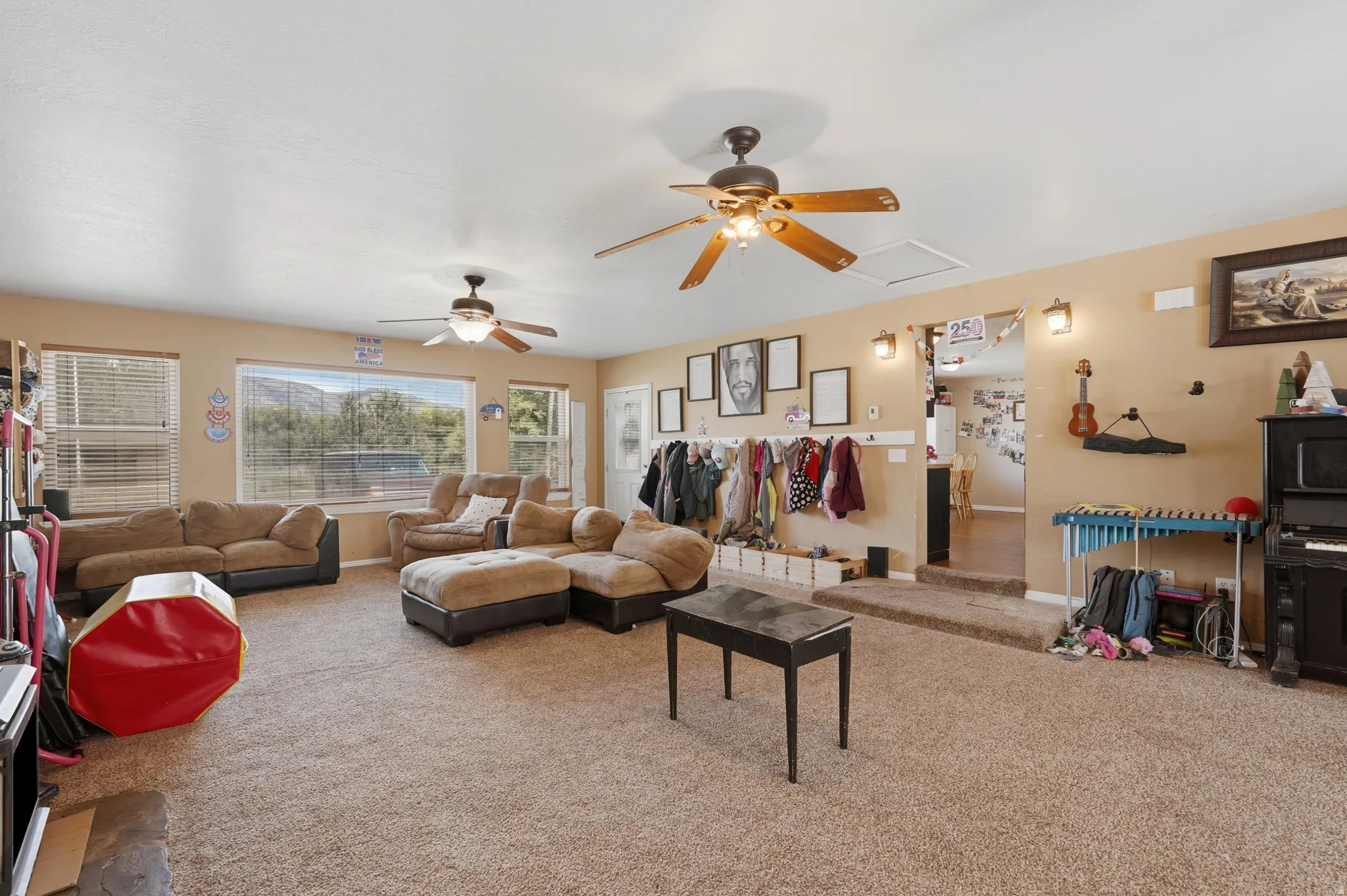 Spacious living area featuring neutral-toned carpeting and walls, multiple windows with horizontal blinds, and two ceiling fans with light fixtures