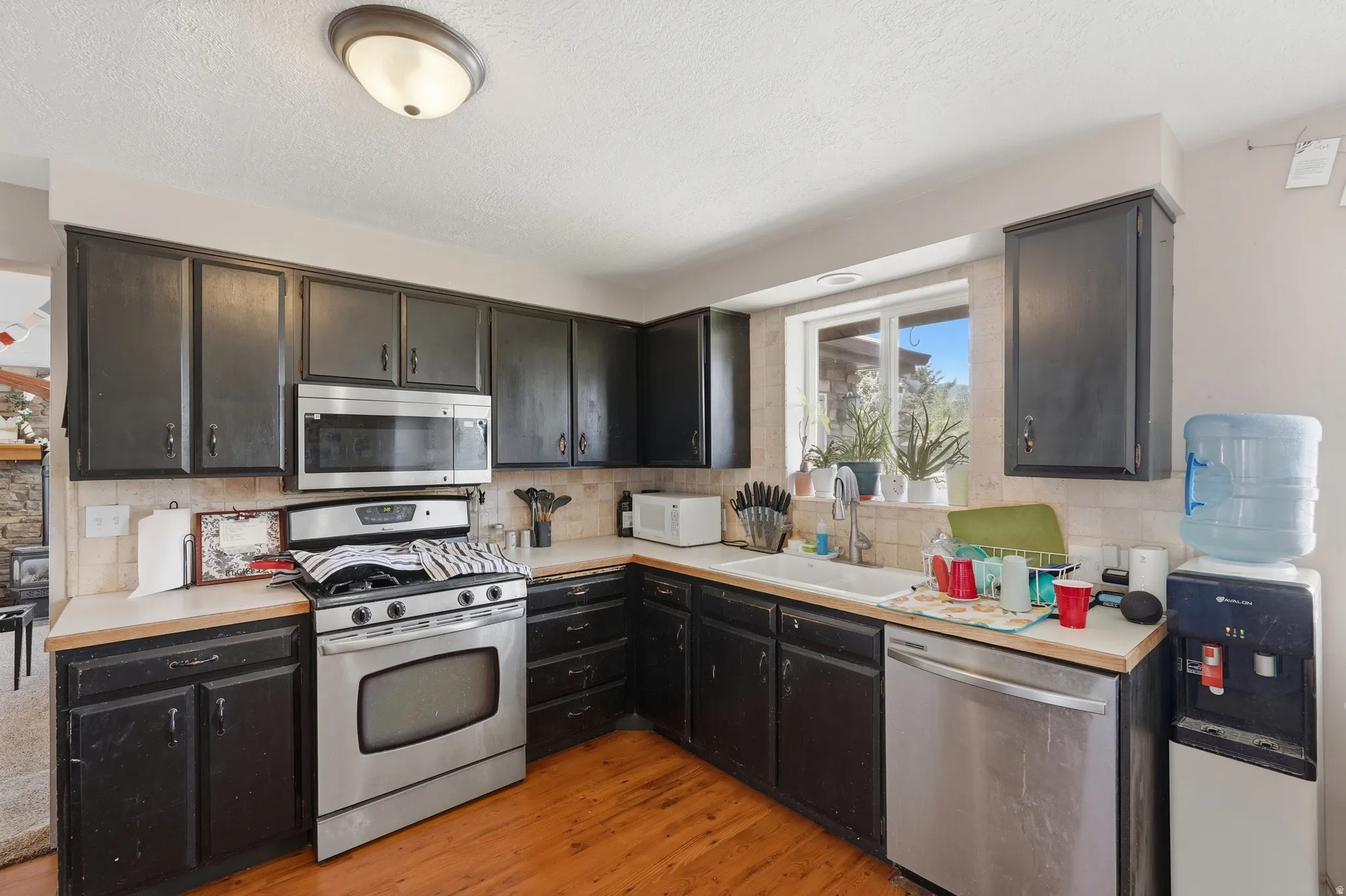 Kitchen featuring black cabinetry, wood-finish flooring, light countertops, stainless steel appliances, and a window above the sink