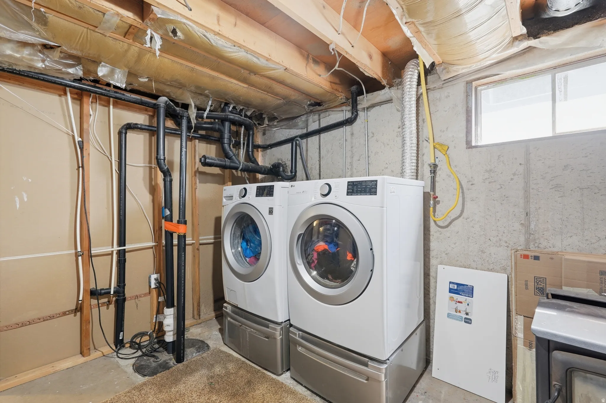 Utility area featuring dual front-loading laundry machines on pedestals, exposed ceiling joists, and concrete walls