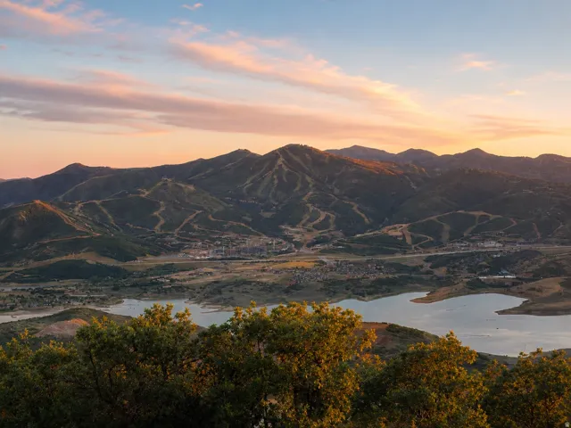 Expansive mountainous landscape featuring a large body of water, distant structures, and visible ski trails