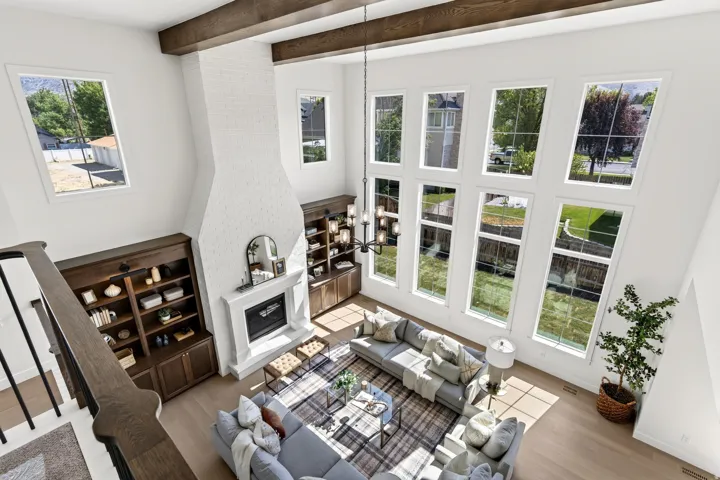 Two-story living area featuring a white brick fireplace with a mantel, built-in shelving, dark wood ceiling beams, and a wall of tall windows